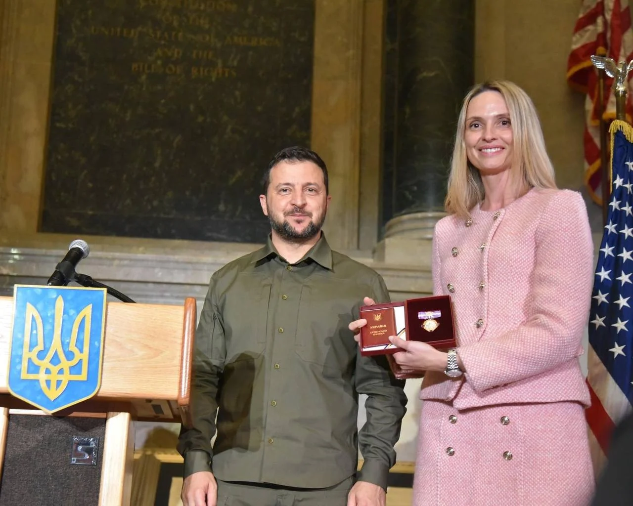 Cérémonie de remise : une femme en veste rose tient une distinction à côté d’un homme en chemise militaire